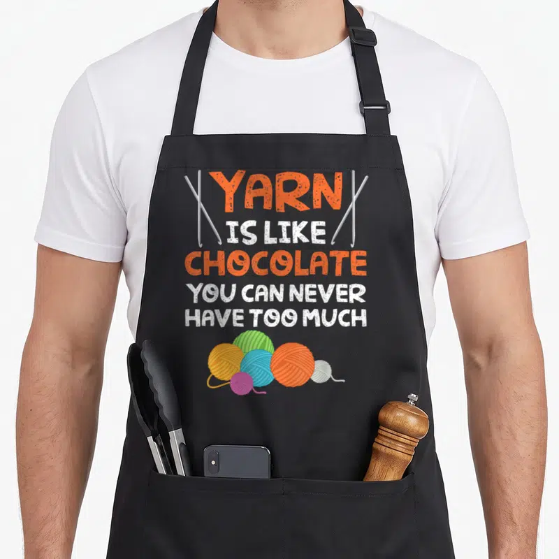 Chocolate knitting apron - Stacked YARN IS LIKE CHOCOLATE text flanked by needles above colorful yarn balls — shown on a black cotton canvas apron against a white background