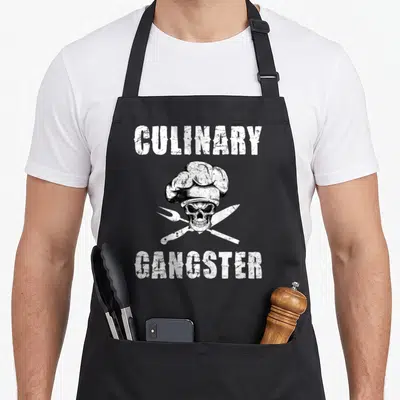 Funny culinary gangster apron - White skull in chef hat with crossed fork knife and distressed text — shown on a black cotton canvas apron against a white background