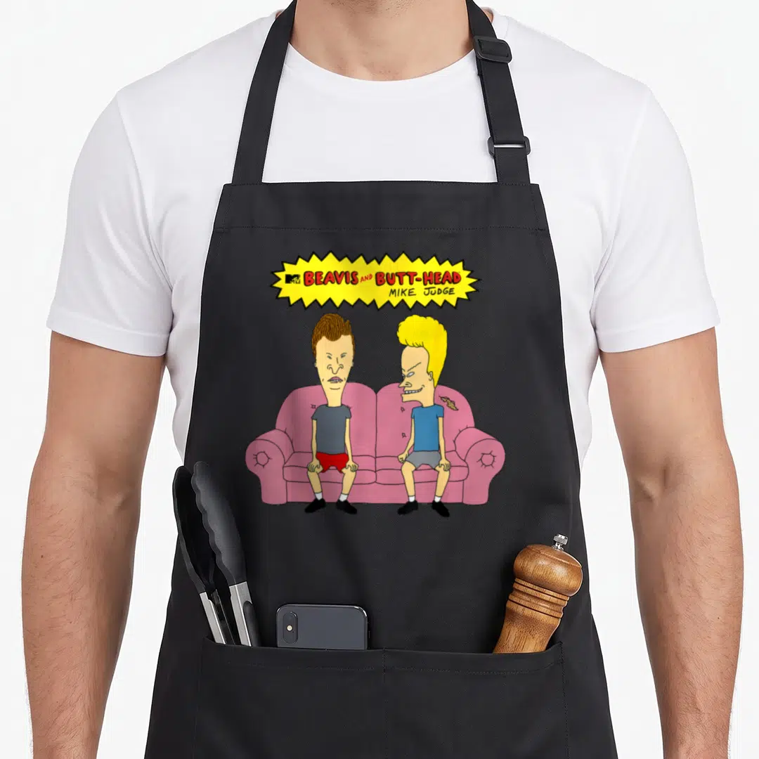 Beavis couch apron - Two cartoon characters on pink couch under yellow burst with red text — shown on a black cotton canvas apron against a white background