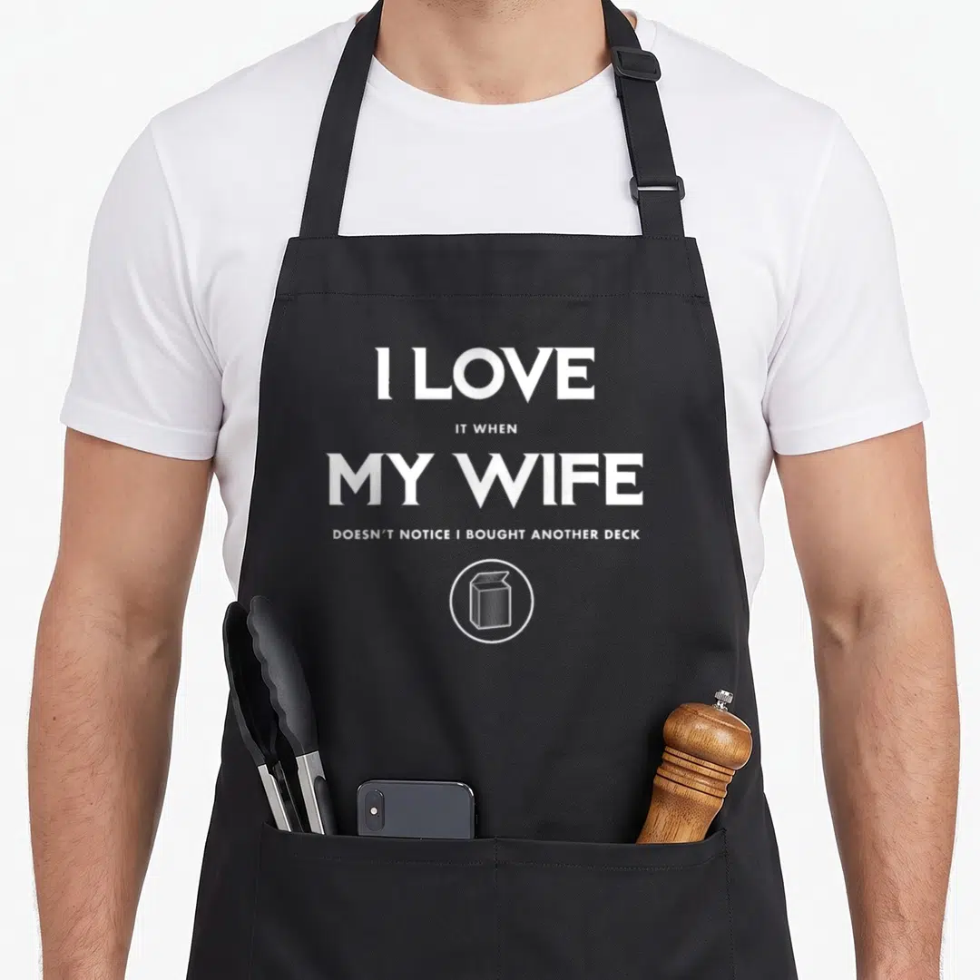 Tcg wife apron - White stacked text reading I LOVE IT WHEN MY WIFE DOESN'T NOTICE I BOUGHT ANOTHER DECK above a deck — shown on a black cotton canvas apron against a white background