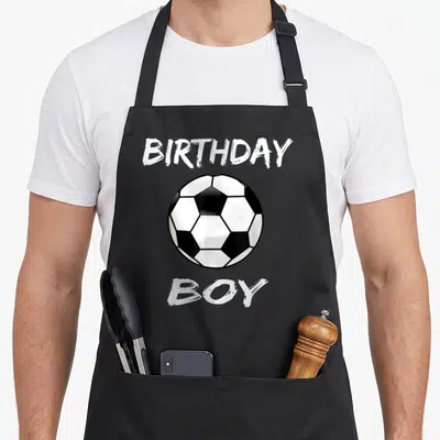 Soccer theme birthday apron - Black and white soccer ball between BIRTHDAY and BOY text in cartoon style — shown on a black cotton canvas apron against a white background