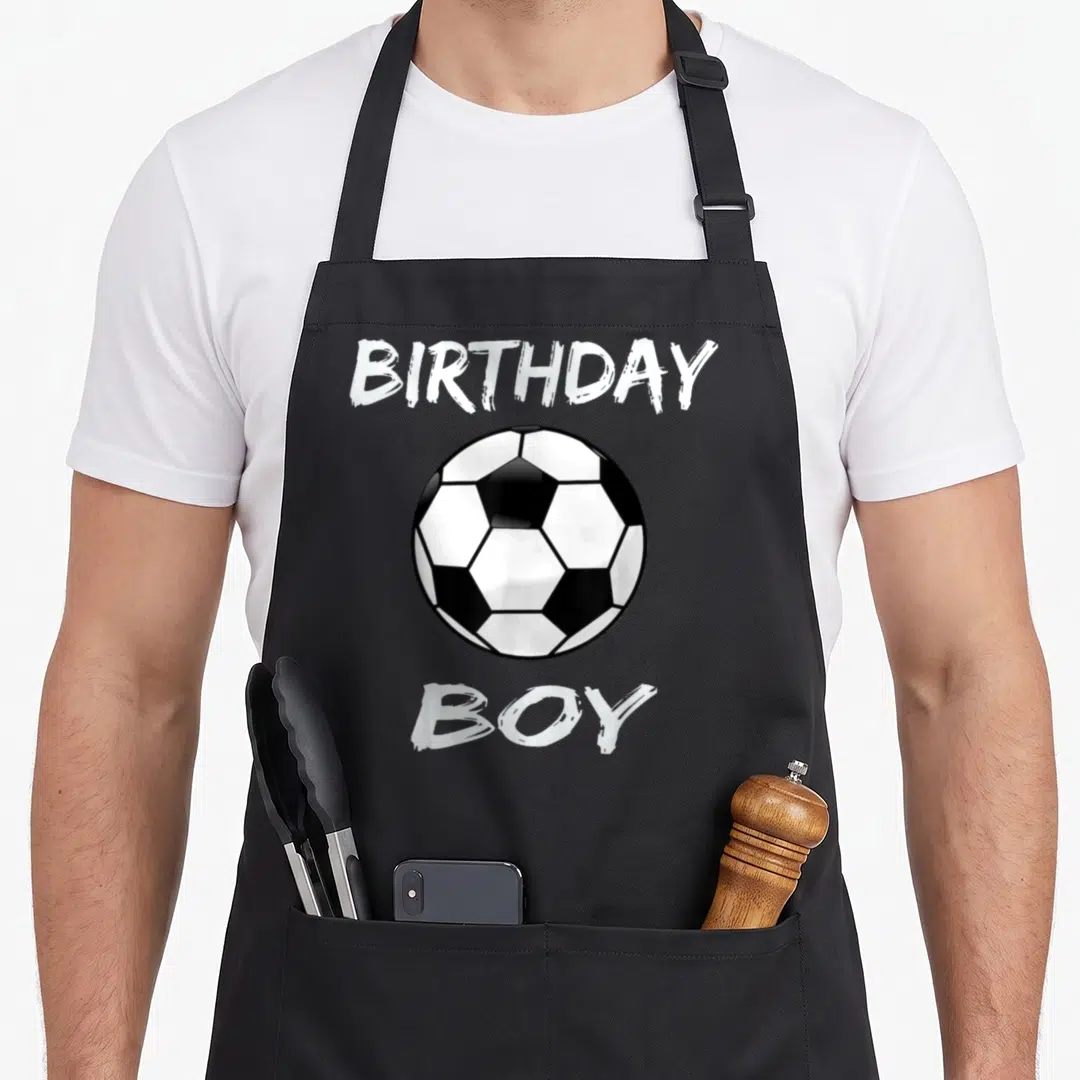 Soccer theme birthday apron - Black and white soccer ball between BIRTHDAY and BOY text in cartoon style — shown on a black cotton canvas apron against a white background