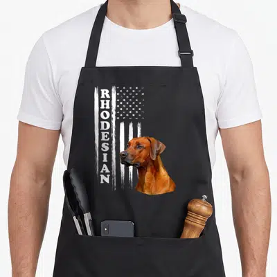 Funny rhodesian ridgeback apron - Realistic brown dog head next to distressed white flag and vertical RHODESIAN text — shown on a black cotton canvas apron against a white background