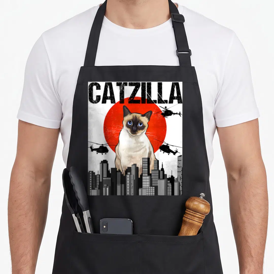 Japanese cute cat aprons - Giant Siamese cat CATZILLA text red sun helicopters city skyline realistic art — shown on a black cotton canvas apron against a white background