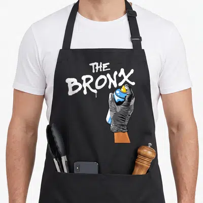 Clothe new york apron - Cartoon hand in black glove holding blue spray can next to white The Bronx graffiti text — shown on a black cotton canvas apron against a white background