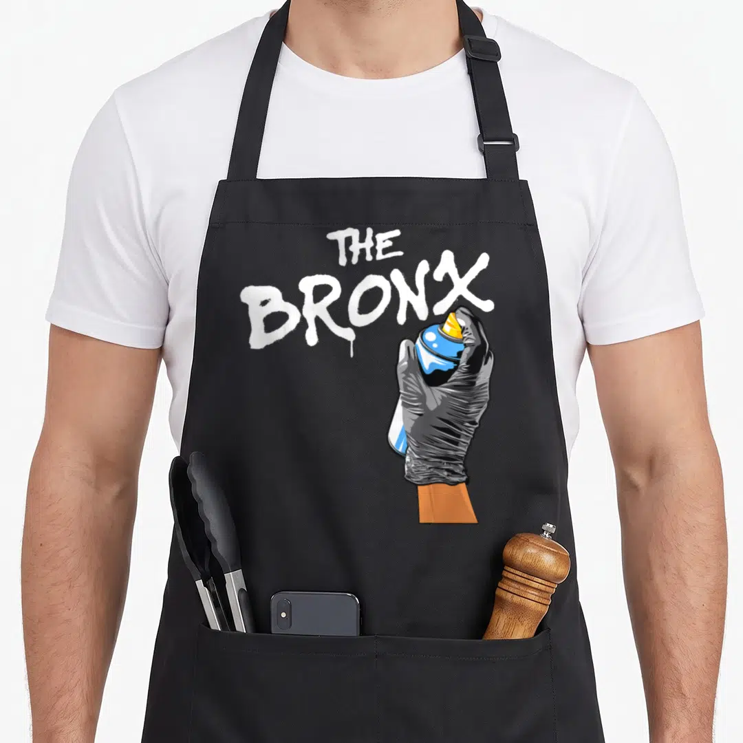 Clothe new york apron - Cartoon hand in black glove holding blue spray can next to white The Bronx graffiti text — shown on a black cotton canvas apron against a white background