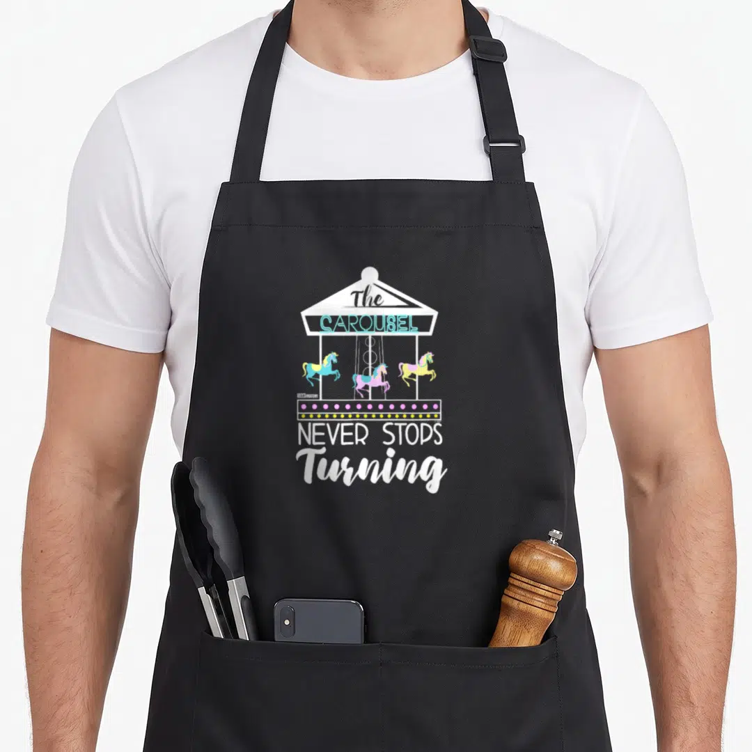 Carousel anatomy apron - Cartoon carousel with three horses and text The CAROUSEL NEVER STOPS Turning — shown on a black cotton canvas apron against a white background