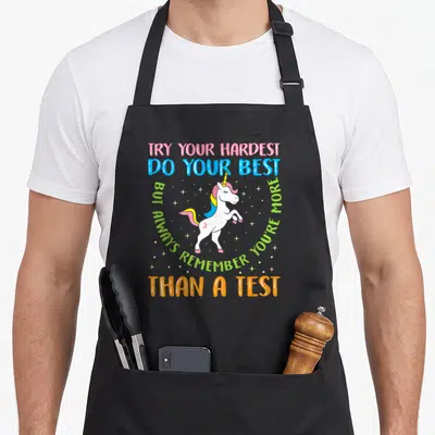 Testing mode teacher apron - Cartoon unicorn with pink mane surrounded by stars and colorful motivational text — shown on a black cotton canvas apron against a white background