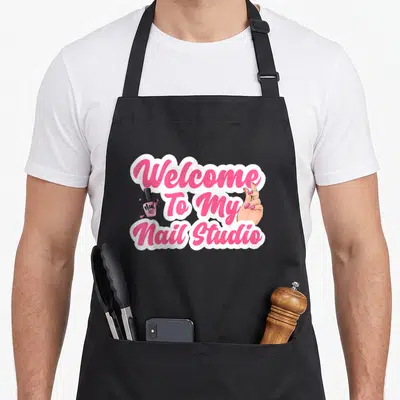 Studio manicurist apron - Pink and white text reading Welcome To My Nail Studio flanked by a polish bottle and hand — shown on a black cotton canvas apron against a white background