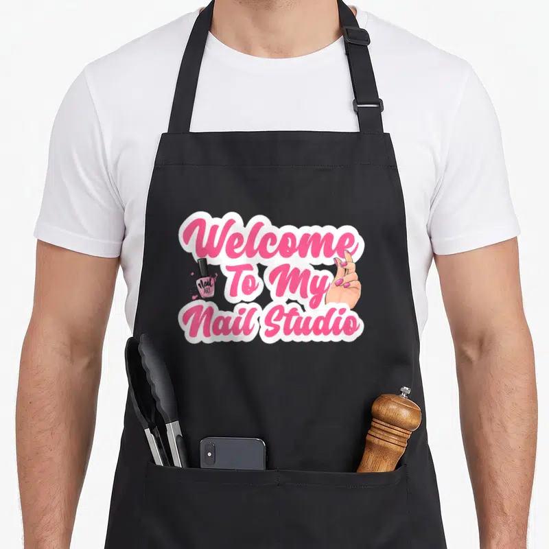 Studio manicurist apron - Pink and white text reading Welcome To My Nail Studio flanked by a polish bottle and hand — shown on a black cotton canvas apron against a white background