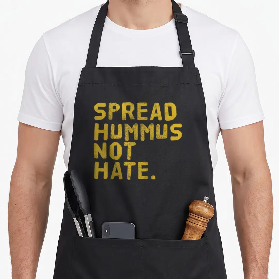 Hummus vegan apron - Yellow stacked text reading SPREAD HUMMUS NOT HATE on black fabric — shown on a black cotton canvas apron against a white background