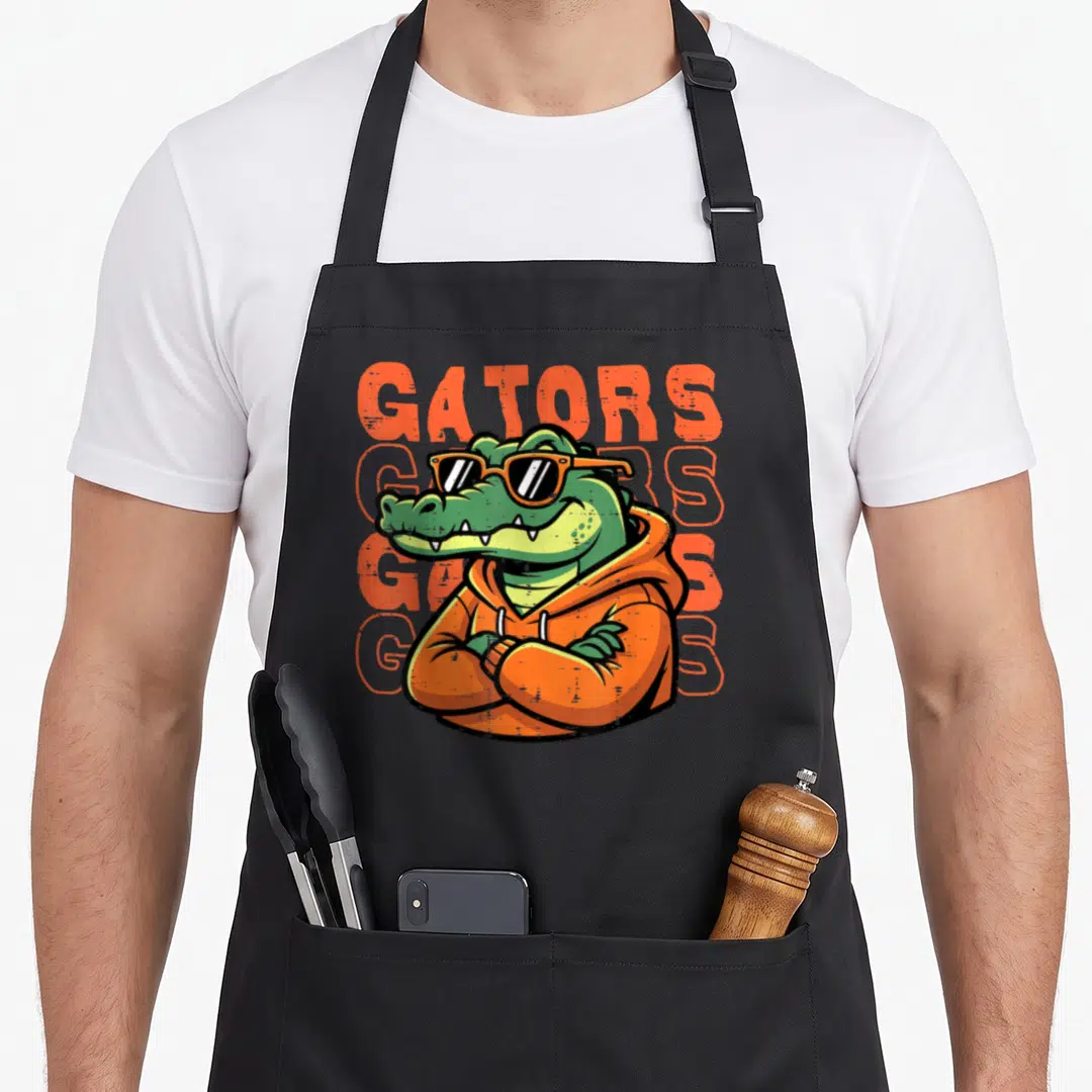 Sunglass gatorade apron - Cartoon alligator in orange hoodie and sunglasses with four GATORS text stacks in orange — shown on a black cotton canvas apron against a white background