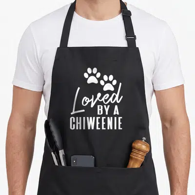 Loved dog apron - Two white paw prints above stacked text reading Loved By A Chiweenie — shown on a black cotton canvas apron against a white background
