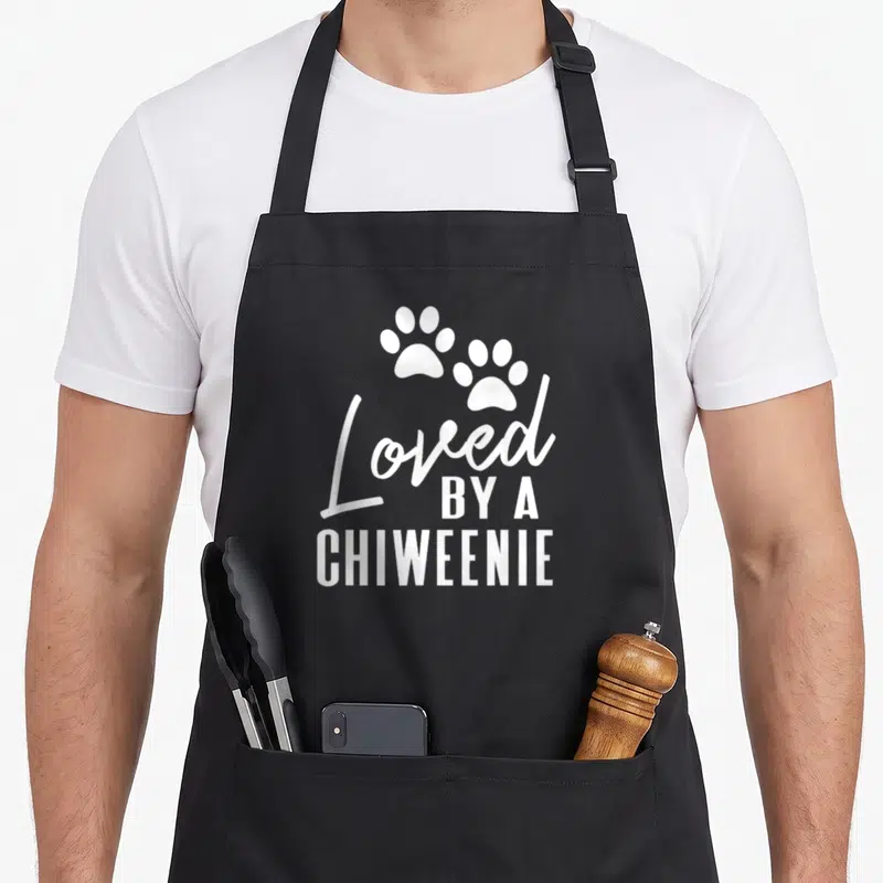 Loved dog apron - Two white paw prints above stacked text reading Loved By A Chiweenie — shown on a black cotton canvas apron against a white background