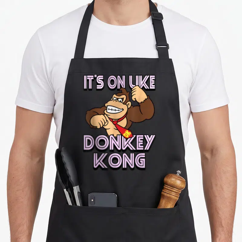 Funny donkey apron - Brown gorilla with red tie and pink It's On Like Donkey Kong text — shown on a black cotton canvas apron against a white background