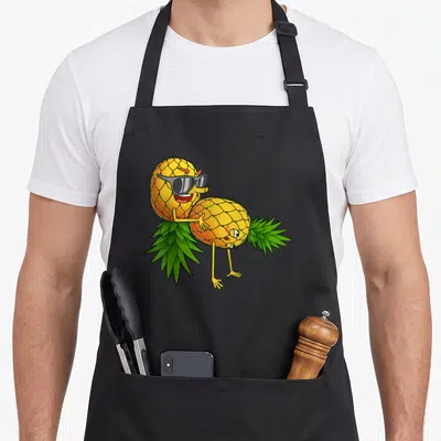 Upside pineapple apron - Two cartoon pineapples hugging, one wearing grey sunglasses, yellow and green art — shown on a black cotton canvas apron against a white background