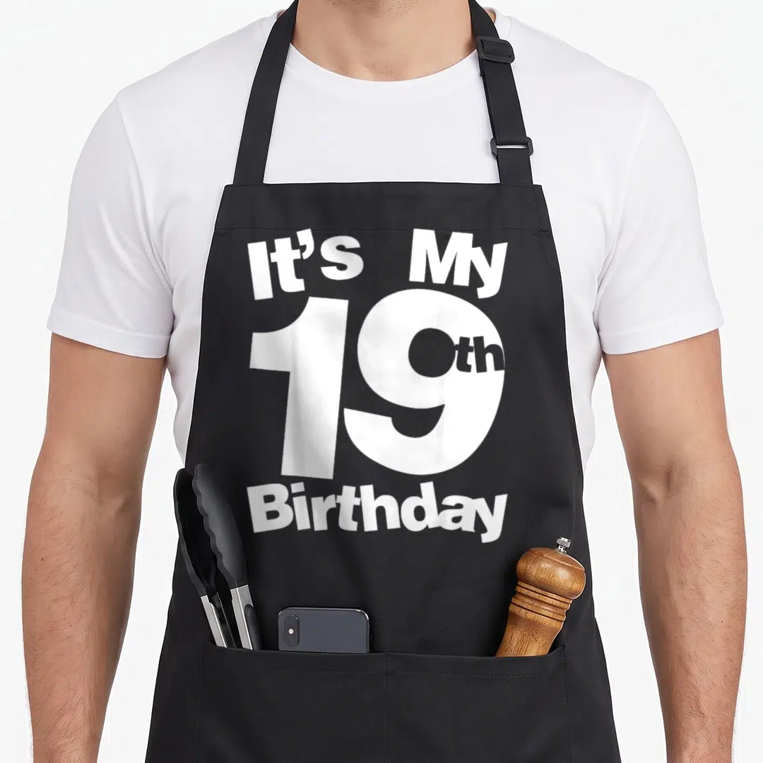 Ist birthday apron - White stacked text reading It's My 19th Birthday on black fabric — shown on a black cotton canvas apron against a white background
