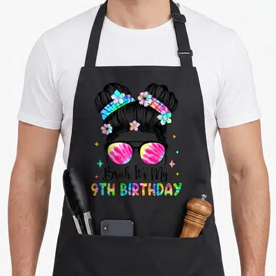 Funny bruh birthday apron - Cartoon girl with space buns and tie-dye sunglasses above 9th Birthday text — shown on a black cotton canvas apron against a white background