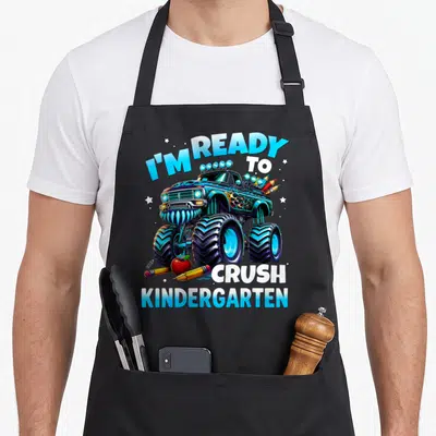 Ready novelty apron - Cartoon monster truck with I'M READY TO CRUSH KINDERGARTEN text, pencils, apple, and stars — shown on a black cotton canvas apron against a white background