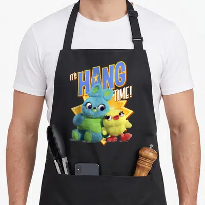 Ducky the story of the apron - Blue plush bunny and chick in front of yellow starburst with IT'S HANG TIME text — shown on a black cotton canvas apron against a white background