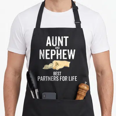 Aunt humorous aprons - White text AUNT AND NEPHEW BEST PARTNERS FOR LIFE above tan cartoon hands pinky swearing — shown on a black cotton canvas apron against a white background