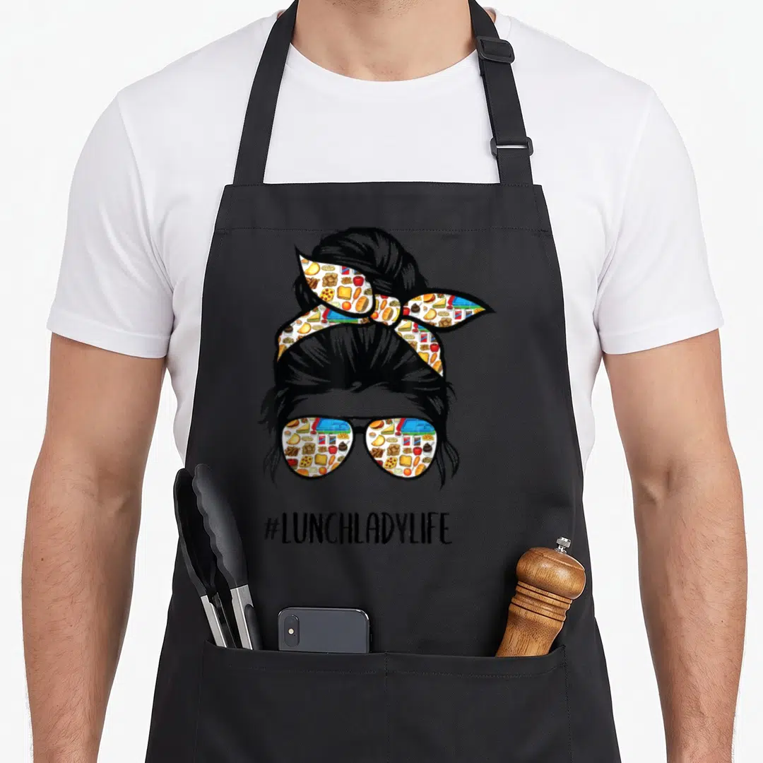 Bun lunch lady apron - Silhouette of woman with bun and food-patterned sunglasses above #LUNCHLADYLIFE text — shown on a black cotton canvas apron against a white background