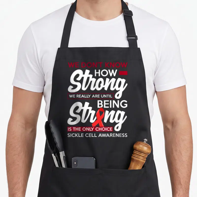 Anemia warrior sickle cell awareness apron - Red and white stacked text reading We Don't Know How Strong with red — shown on a black cotton canvas apron against a white background