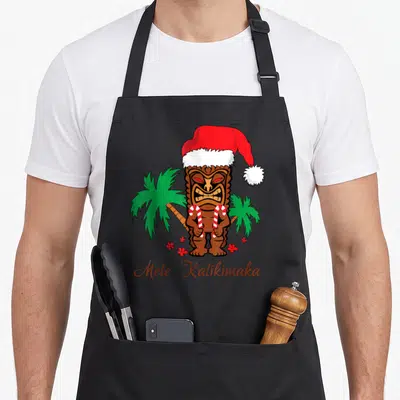 Mele merry christmas apron - Brown tiki statue in red santa hat holding two candy canes between green palms with red — shown on a black cotton canvas apron against a white background