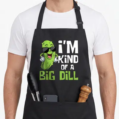 Kind vegetable apron - Cartoon pickle with sunglasses next to stacked text reading I'M KIND OF A BIG DILL — shown on a black cotton canvas apron against a white background