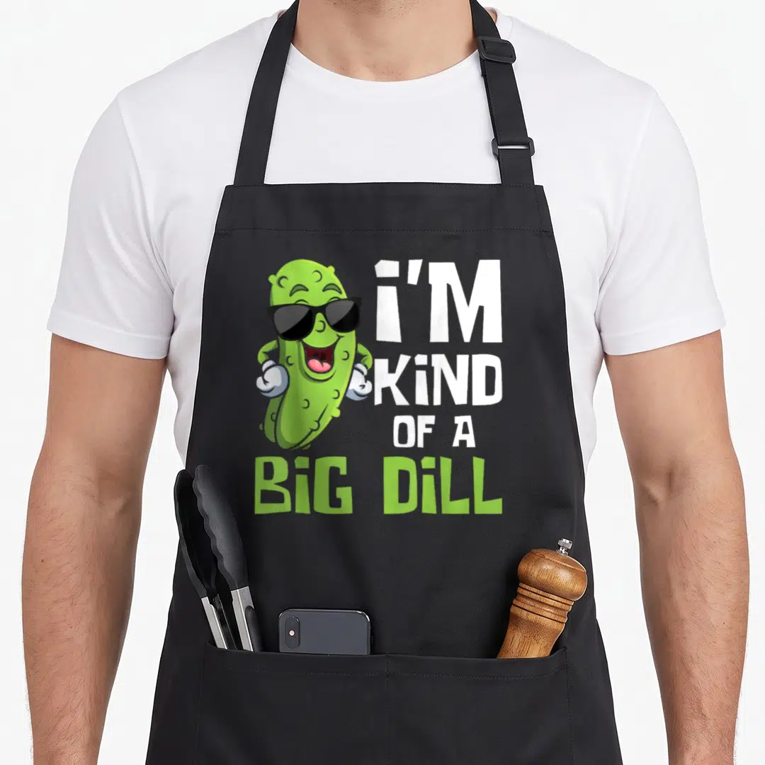 Kind vegetable apron - Cartoon pickle with sunglasses next to stacked text reading I'M KIND OF A BIG DILL — shown on a black cotton canvas apron against a white background