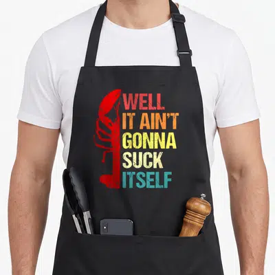 Well cajun aprons - Red lobster silhouette beside stacked multi-colored text reading Well It Ain't Gonna Suck Itself — shown on a black cotton canvas apron against a white background