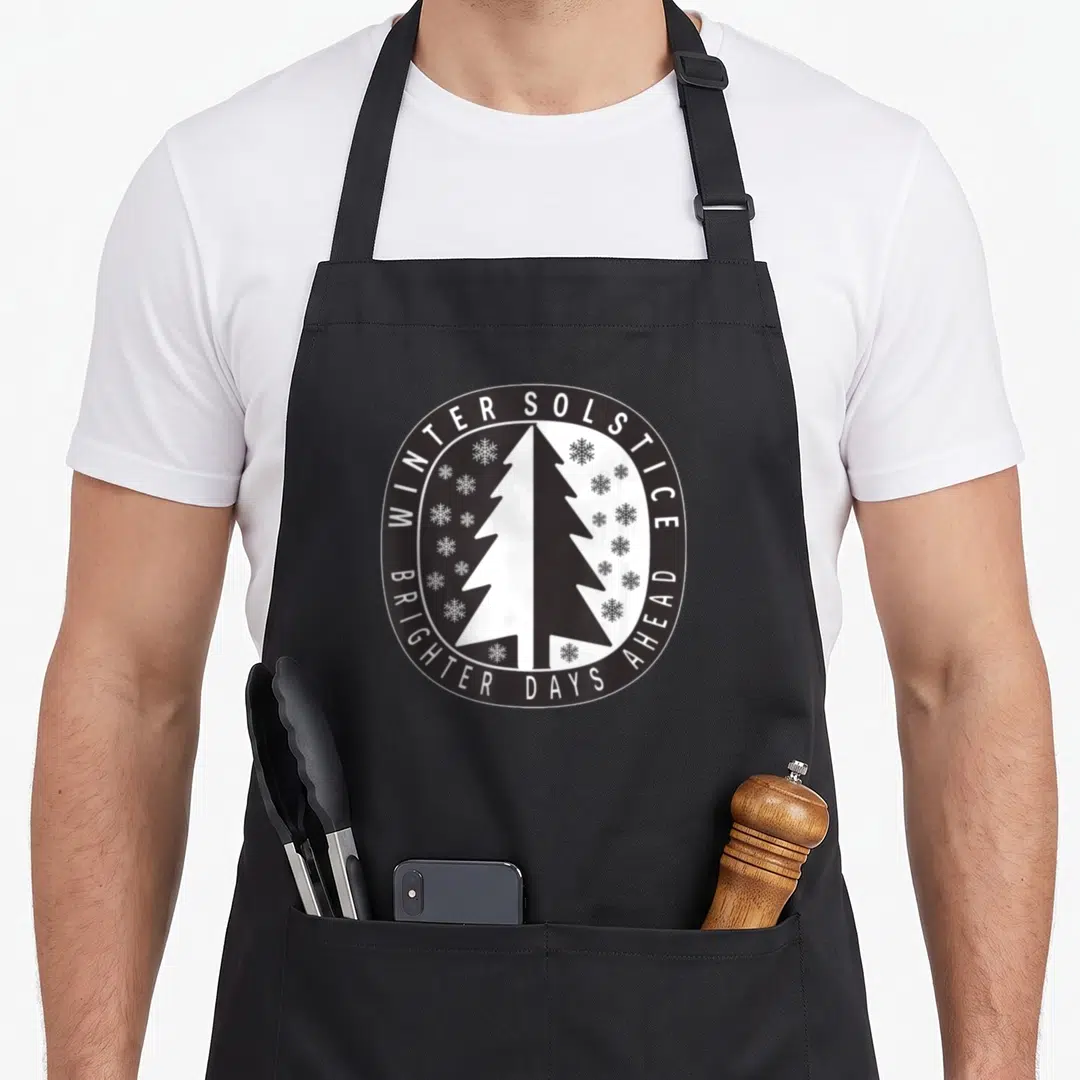 Brighter winter apron - Circular black and white design with split tree and snowflakes reading Winter Solstice Brighter — shown on a black cotton canvas apron against a white background