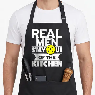 Stay paddleball pickleball apron - Yellow perforated ball overlaid on stacked white text reading REAL MEN STAY OUT — shown on a black cotton canvas apron against a white background