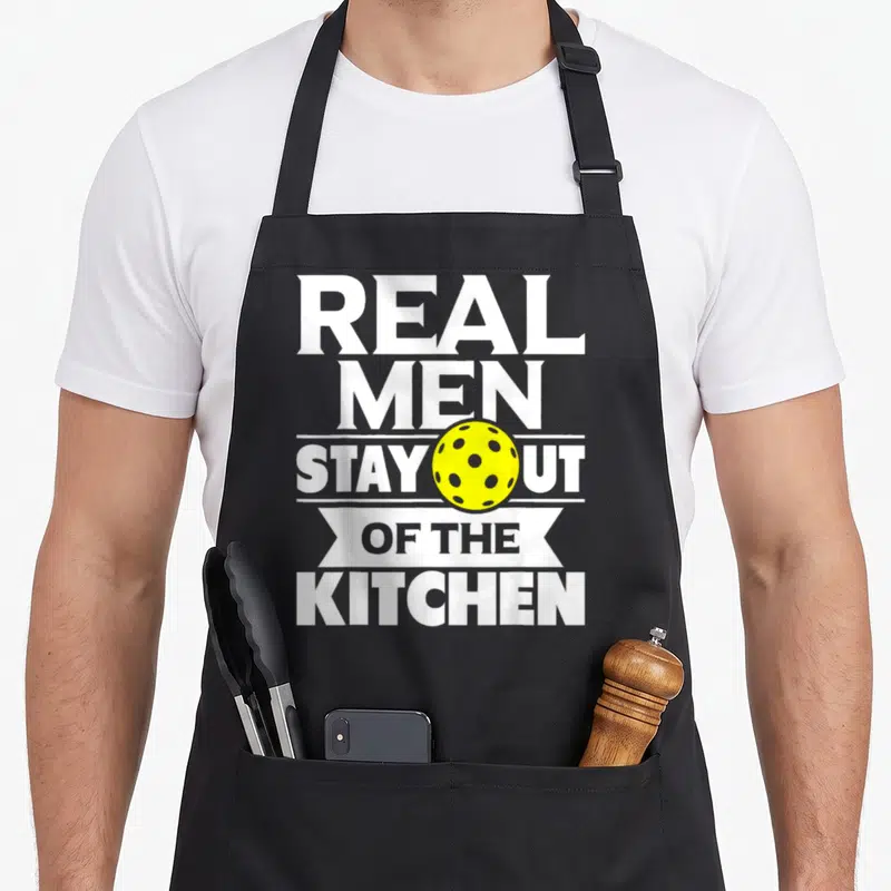 Stay paddleball pickleball apron - Yellow perforated ball overlaid on stacked white text reading REAL MEN STAY OUT — shown on a black cotton canvas apron against a white background