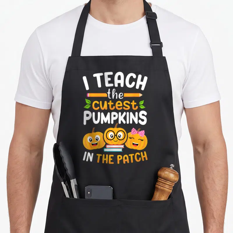 Teach halloween kitchen aprons - Stacked text I TEACH THE CUTEST PUMPKINS IN THE PATCH with three cartoon pumpkins — shown on a black cotton canvas apron against a white background
