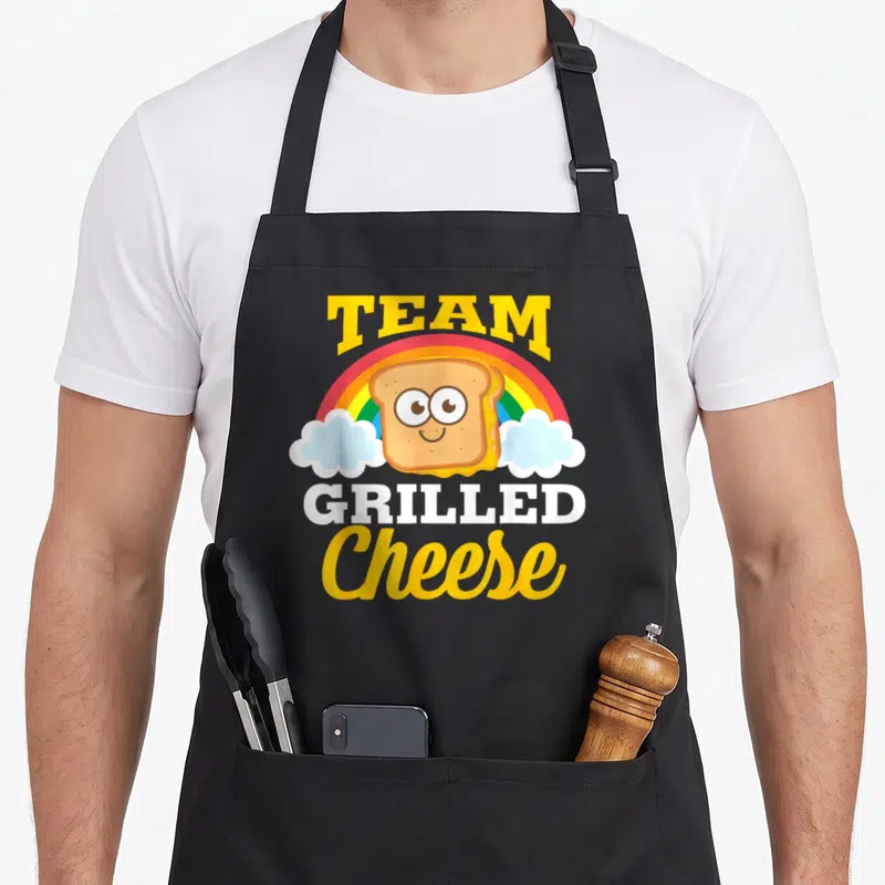 Cheddar cheese apron - Cartoon smiling grilled sandwich with rainbow, clouds, and TEAM GRILLED Cheese text in yellow — shown on a black cotton canvas apron against a white background
