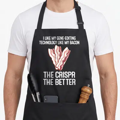 Crispr science apron - White text reading I like my gene-editing technology like my bacon the crispr the better around — shown on a black cotton canvas apron against a white background