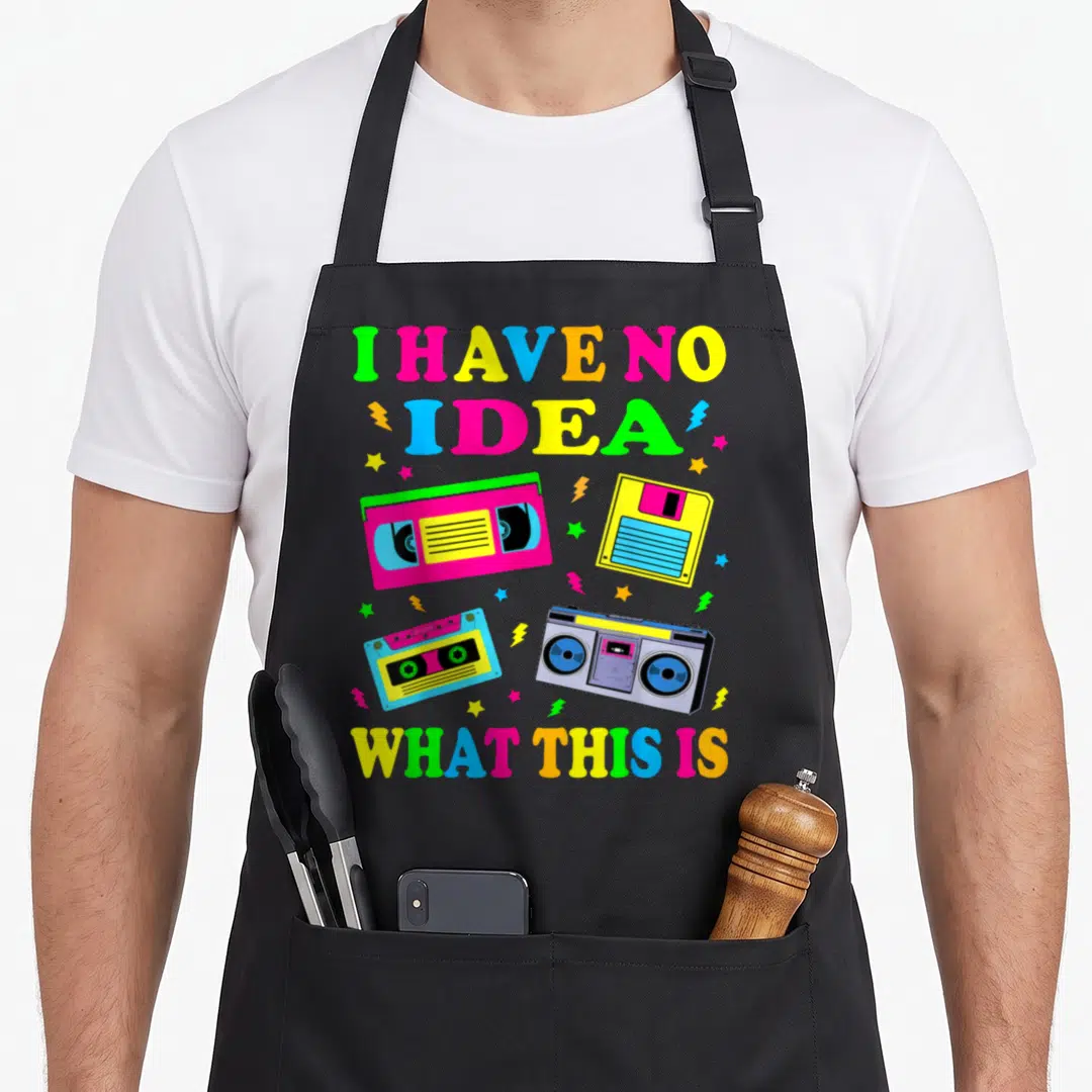 70s chef outfit with apron - I HAVE NO IDEA WHAT THIS IS text with pink VHS, yellow cassette, and blue boombox graphics — shown on a black cotton canvas apron against a white background