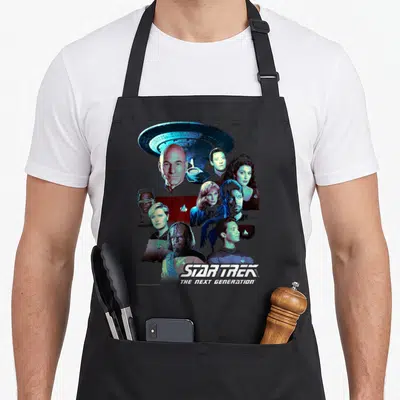 Generation crew apron - Star Trek Next Generation cast collage below spaceship with blue white red uniforms — shown on a black cotton canvas apron against a white background