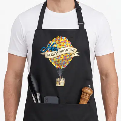 Greatest hitching up apron - House suspended under multicolored balloons with beige ribbon reading Her GREATEST — shown on a black cotton canvas apron against a white background