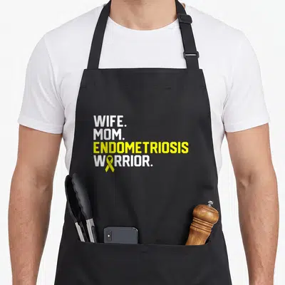 Well wife apron - WIFE MOM ENDOMETRIOSIS WARRIOR text with yellow ribbon over O — shown on a black cotton canvas apron against a white background