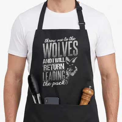 Throw wolves apron - Stacked white distressed text reading throw me to the WOLVES AND I WILL RETURN LEADING the pack — shown on a black cotton canvas apron against a white background