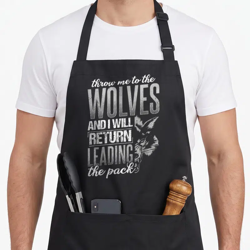 Throw wolves apron - Stacked white distressed text reading throw me to the WOLVES AND I WILL RETURN LEADING the pack — shown on a black cotton canvas apron against a white background