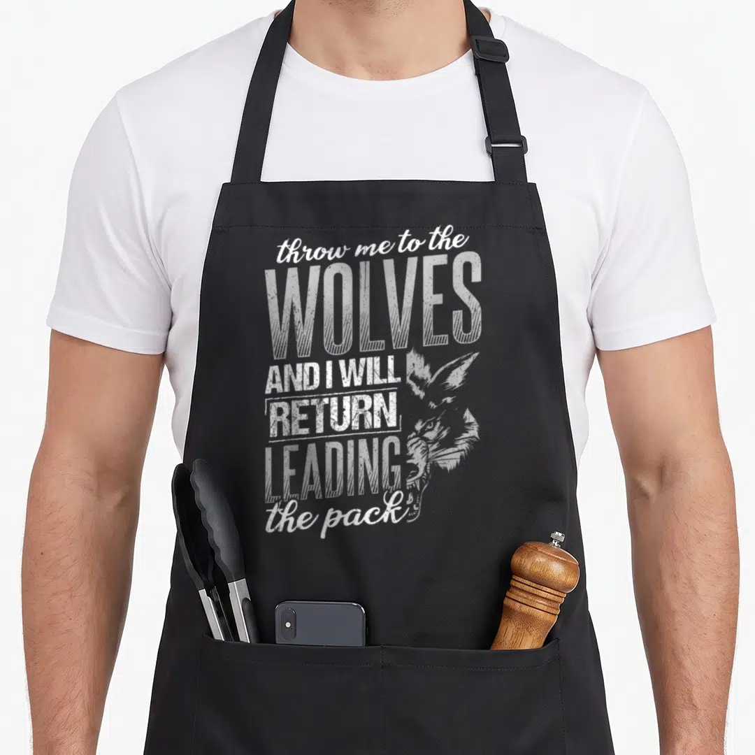Throw wolves apron - Stacked white distressed text reading throw me to the WOLVES AND I WILL RETURN LEADING the pack — shown on a black cotton canvas apron against a white background