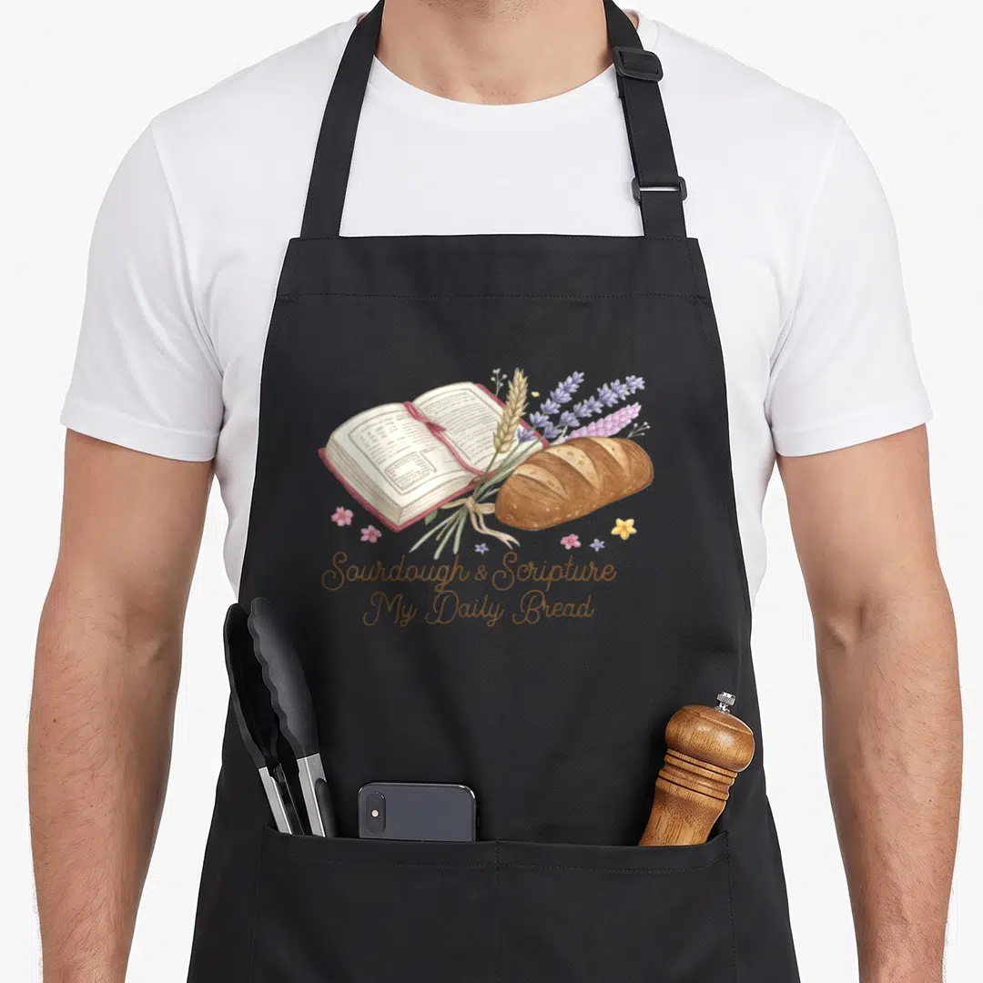Scripture sourdough bread apron - Open bible, brown loaf, purple lavender, and brown script text in watercolor style — shown on a black cotton canvas apron against a white background