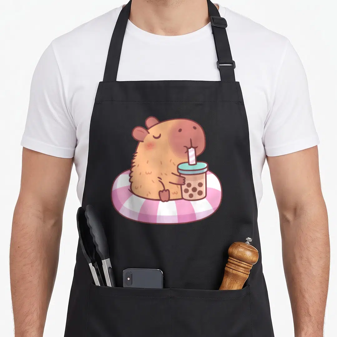 Pool anime apron - Brown capybara in pink white swim ring drinking bubble tea cartoon — shown on a black cotton canvas apron against a white background