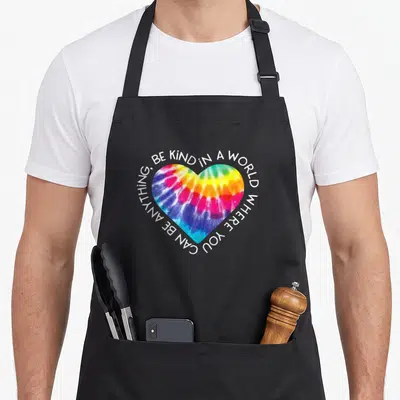 Anti tie-dye apron - Multicolored heart with circular white text reading Be Kind In A World Where You Can Be Anything — shown on a black cotton canvas apron against a white background