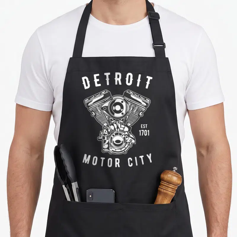 Detroit block & apron - V-twin engine illustration with Detroit Est 1701 Motor City text in distressed white and grey — shown on a black cotton canvas apron against a white background