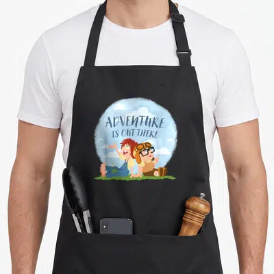 Funny adventure carl and ellie apron - Two children on green grass under blue sky with text ADVENTURE IS OUT THERE — shown on a black cotton canvas apron against a white background