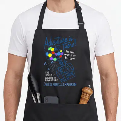 Cute adventure balloon apron - Blue line-art house floating on colorful balloons with text reading Adventure is out — shown on a black cotton canvas apron against a white background
